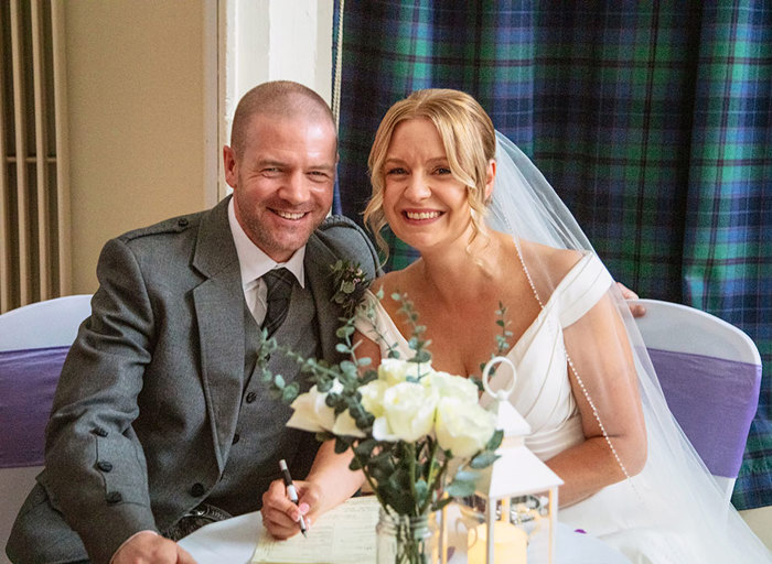 A man in a grey kilt and a woman in a wedding dress and veil sign their marriage license 