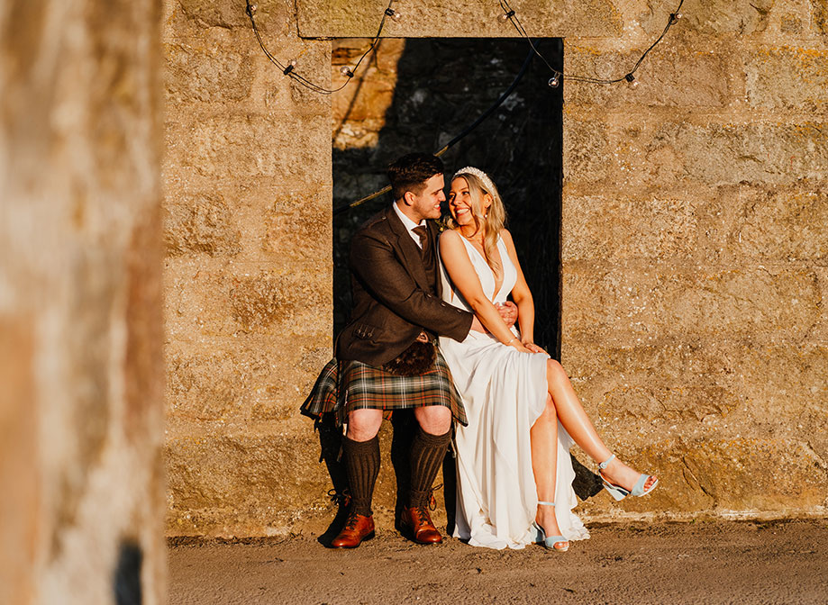 a bride and groom bathed in golden sunlight. They are sitting on an alcove within a stone wall