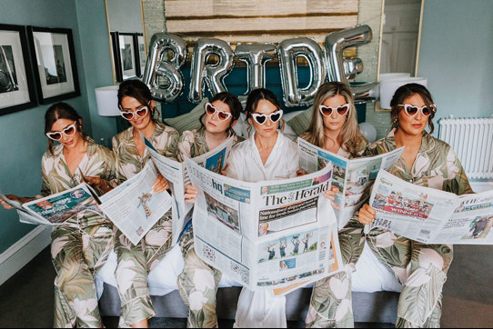 a bride and her bridesmaids in pajamas and sunglasses while reading newspapers 