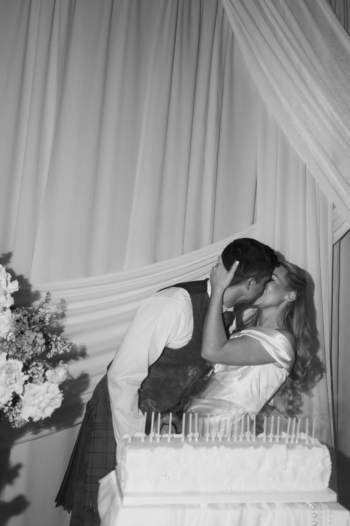 Siân and Stuart kiss behind their long rectangular wedding cake, handmade by the bride’s sister, at their Mar Hall reception.