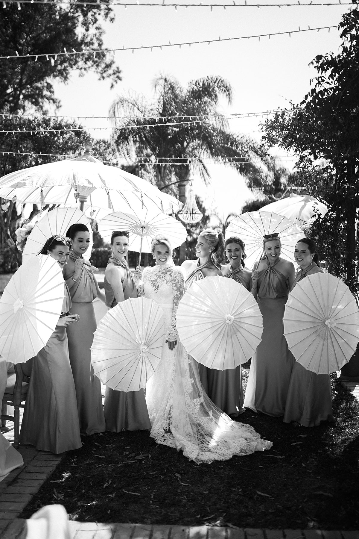 Bride and bridesmaids pose outdoors holding white paper parasols at a summer wedding