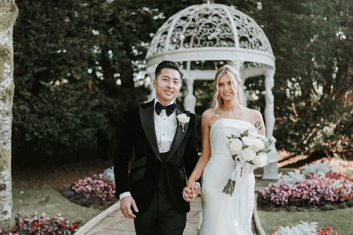 a bride and groom walking hand in hand in garden at busby hotel with white pergola in background