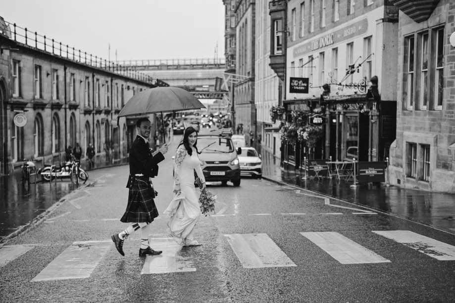Black and white photo of groom holding umbrella over bride as they walk through Edinburgh