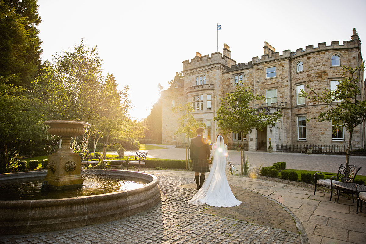 bride and groom walk hand in hand towards crossbasket castle as the sun sets in the background