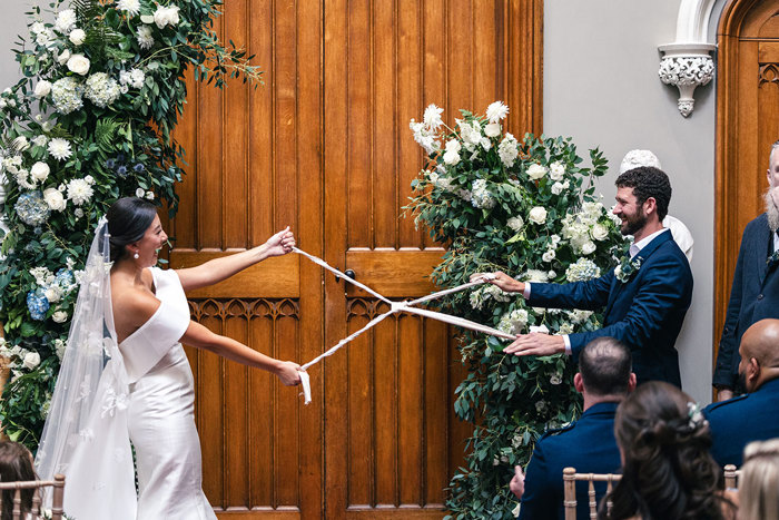 An elated bride and groom tying the knot during a handfasting ritual while standing near a large warm wooden door that's flanked by an asymmetric abundant white and blue flower floor-standing decoration