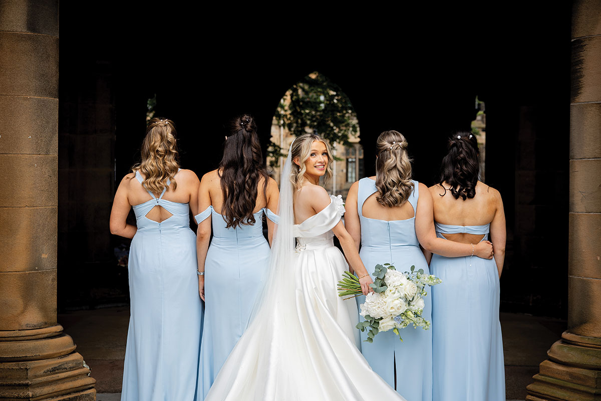 Bride in a white gown standing with bridesmaids in pale blue dresses beneath a stone archway, looking back towards the camera