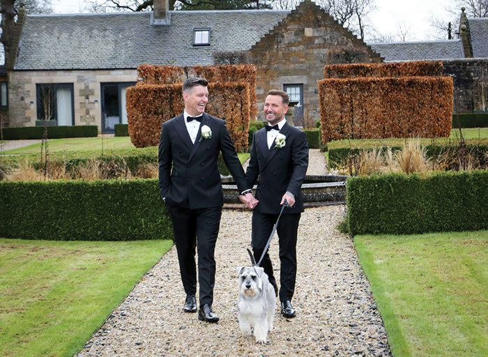 two grooms walking along a gravel path in the gardens at Carlowrie Castle with a schnauzer dog on a lead. There is a building in the background
