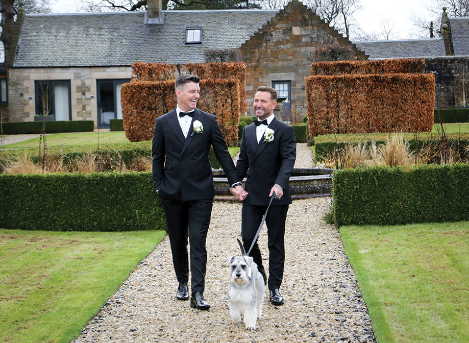 two grooms walking along a gravel path in the gardens at Carlowrie Castle with a schnauzer dog on a lead. There is a building in the background