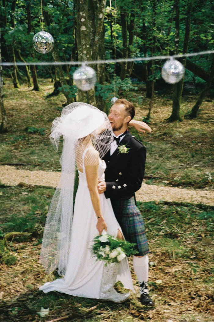 a bride wearing a large white hat with netting and groom wearing a kilt standing in a wooded setting.