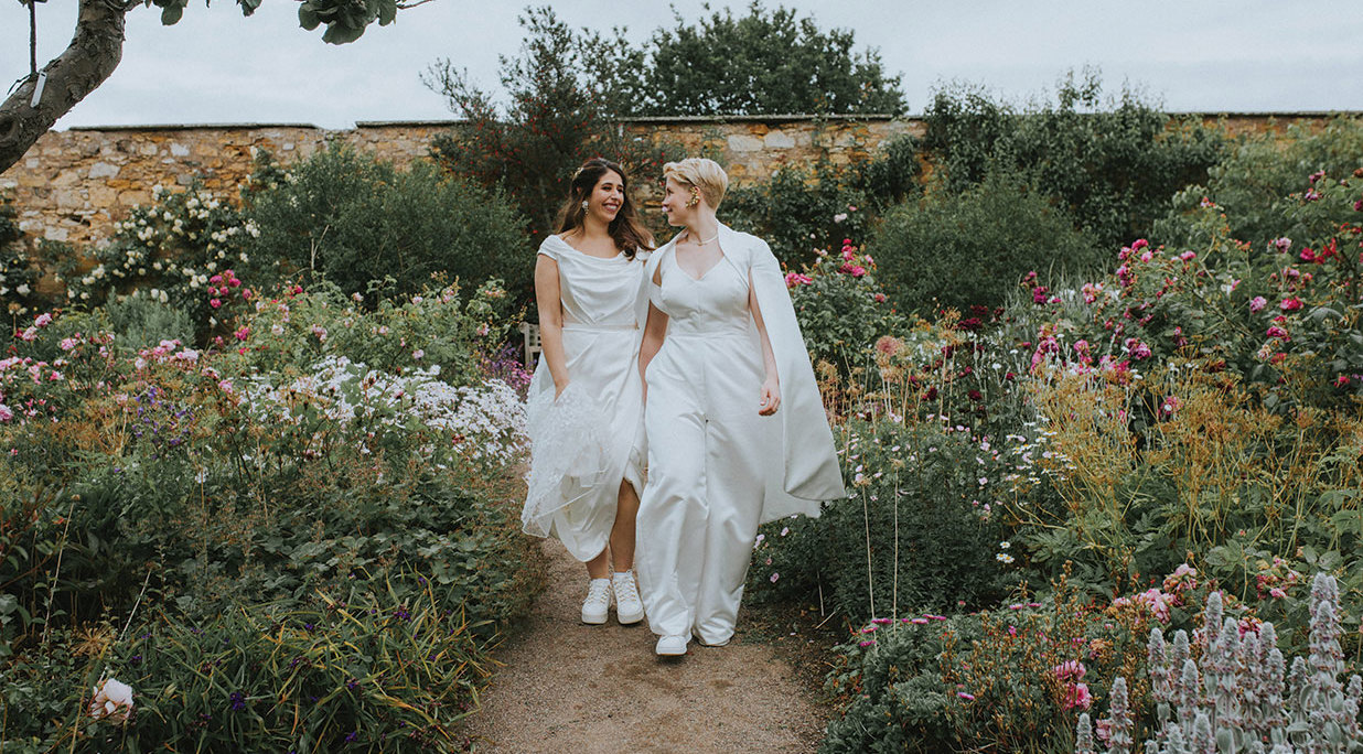 two brides walking hand-in-hand down a path in a walled garden filled with flowers at Cambo House Fife wedding venue