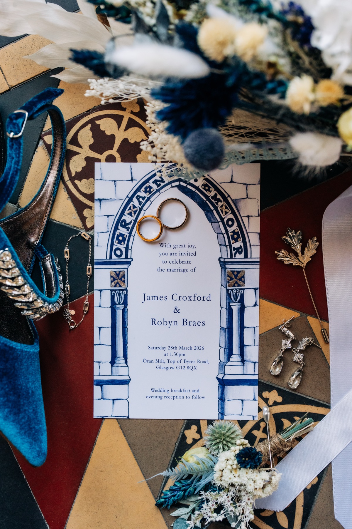 Flat lay of Robyn and James's Òran Mór wedding invitation, rings, blue bridal shoes, jewellery and flowers arranged on patterned tiles.