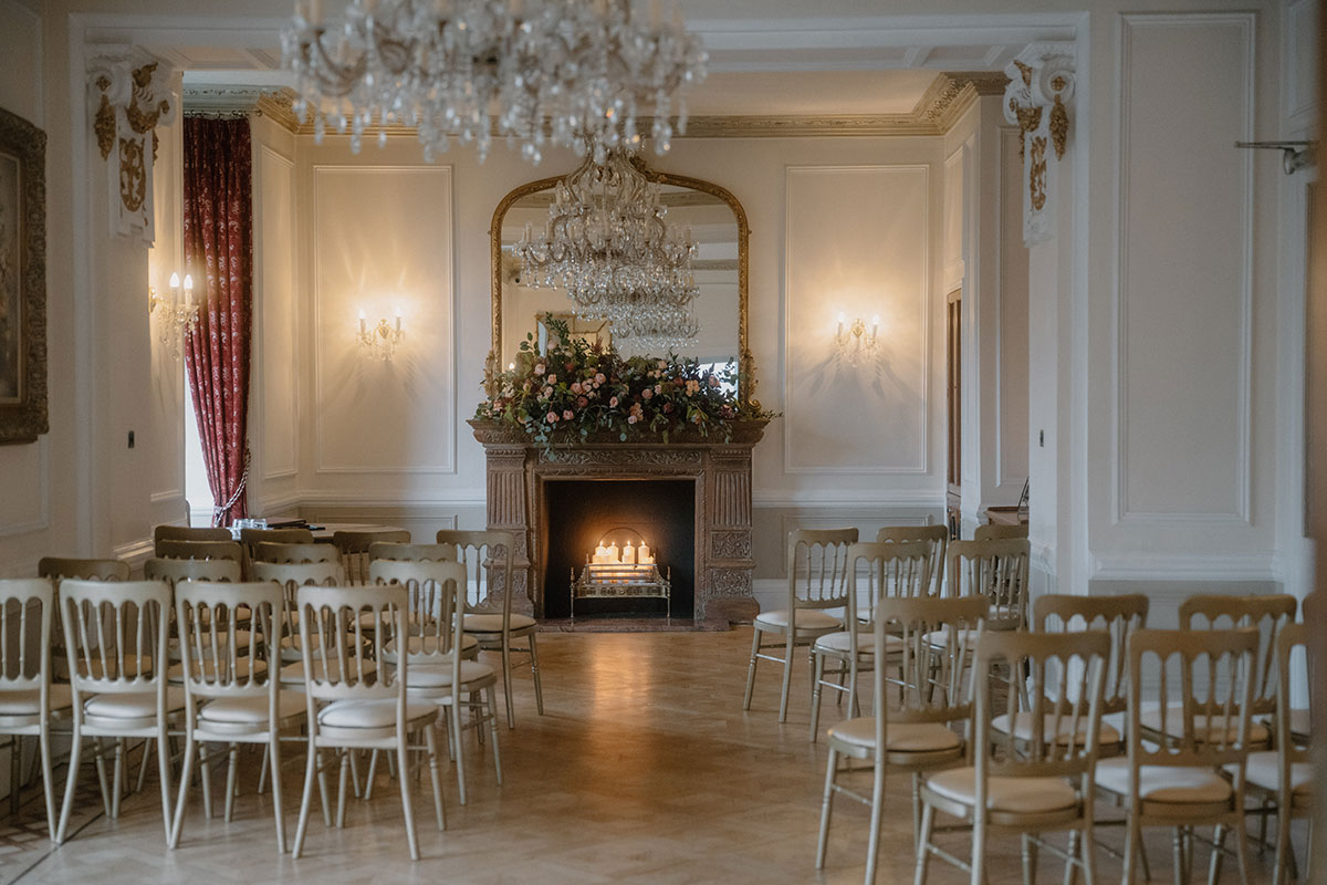 Ceremony room at Auchen Castle arranged with cream chairs facing a candlelit fireplace adorned with floral arrangements beneath crystal chandeliers