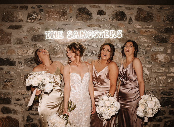 a joyful bride and three bridesmaids wearing satin dresses stand against a rustic brick wall below a neon sign
