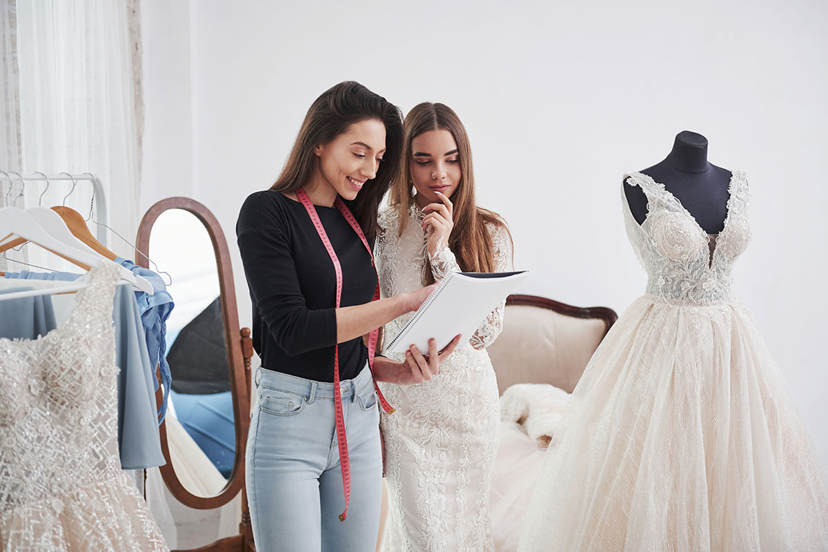 dark brunette woman with pink measuring tape hanging around her neck holds notebook and chats to another brunette woman in a wedding dress, pointing at the notebook as they stand in a changing room with other wedding dresses on display