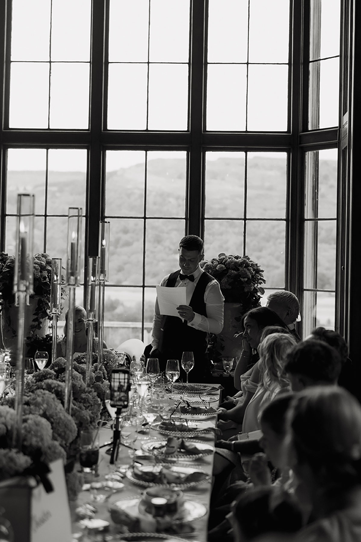 Groom delivering his wedding speech beside a long candlelit table with views of the hills through large windows.