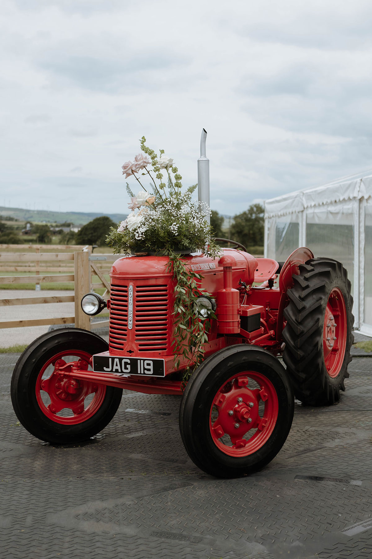 Red vintage tractor decorated with wedding flowers and greenery outside a marquee venue.