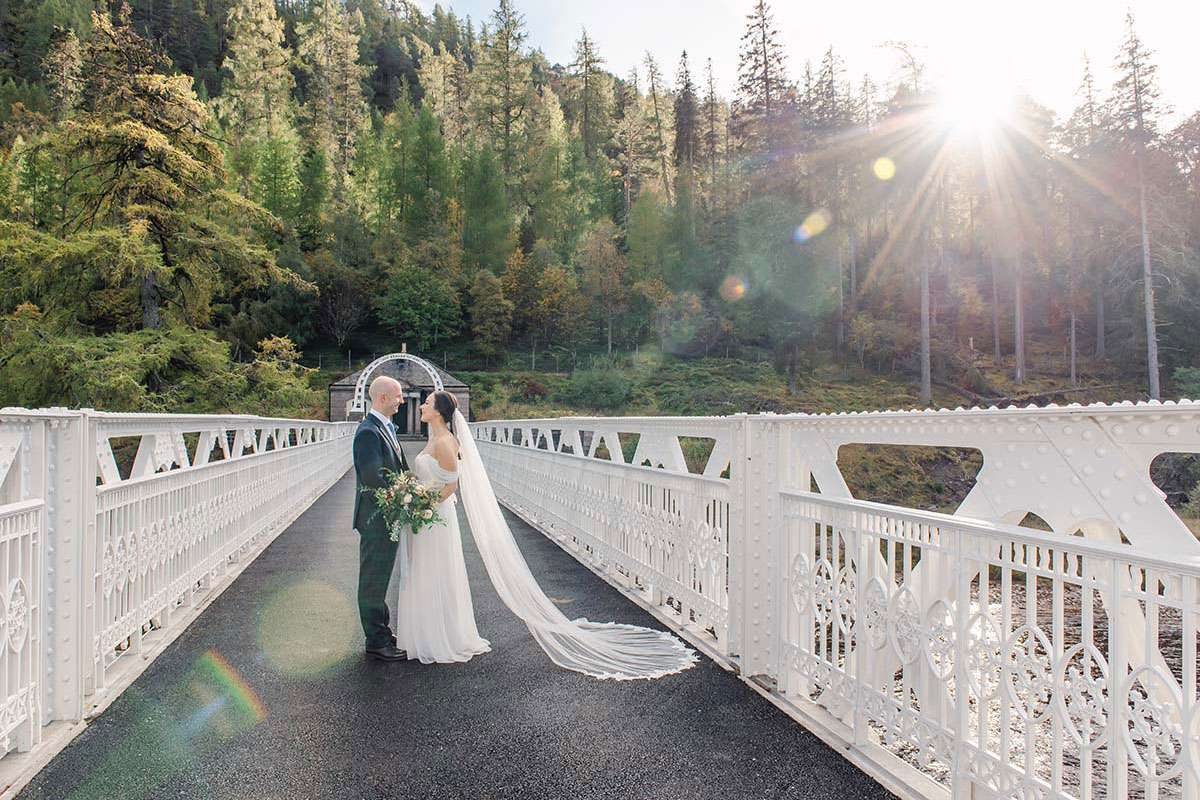 Bride and groom facing each other on a white metal bridge surrounded by forest, with sunlight streaming through the trees