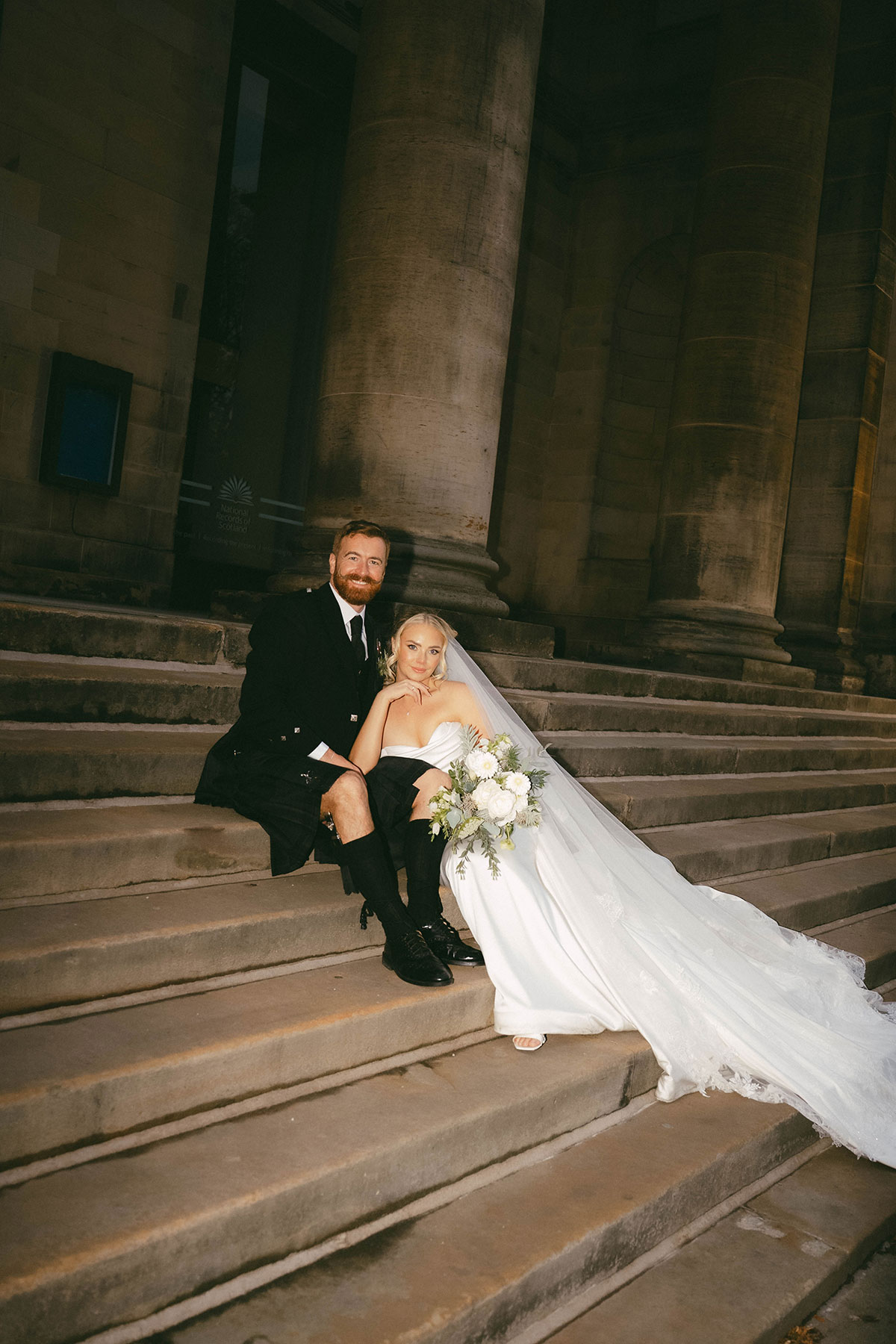 bride and groom sitting on stone steps outside historic building at night