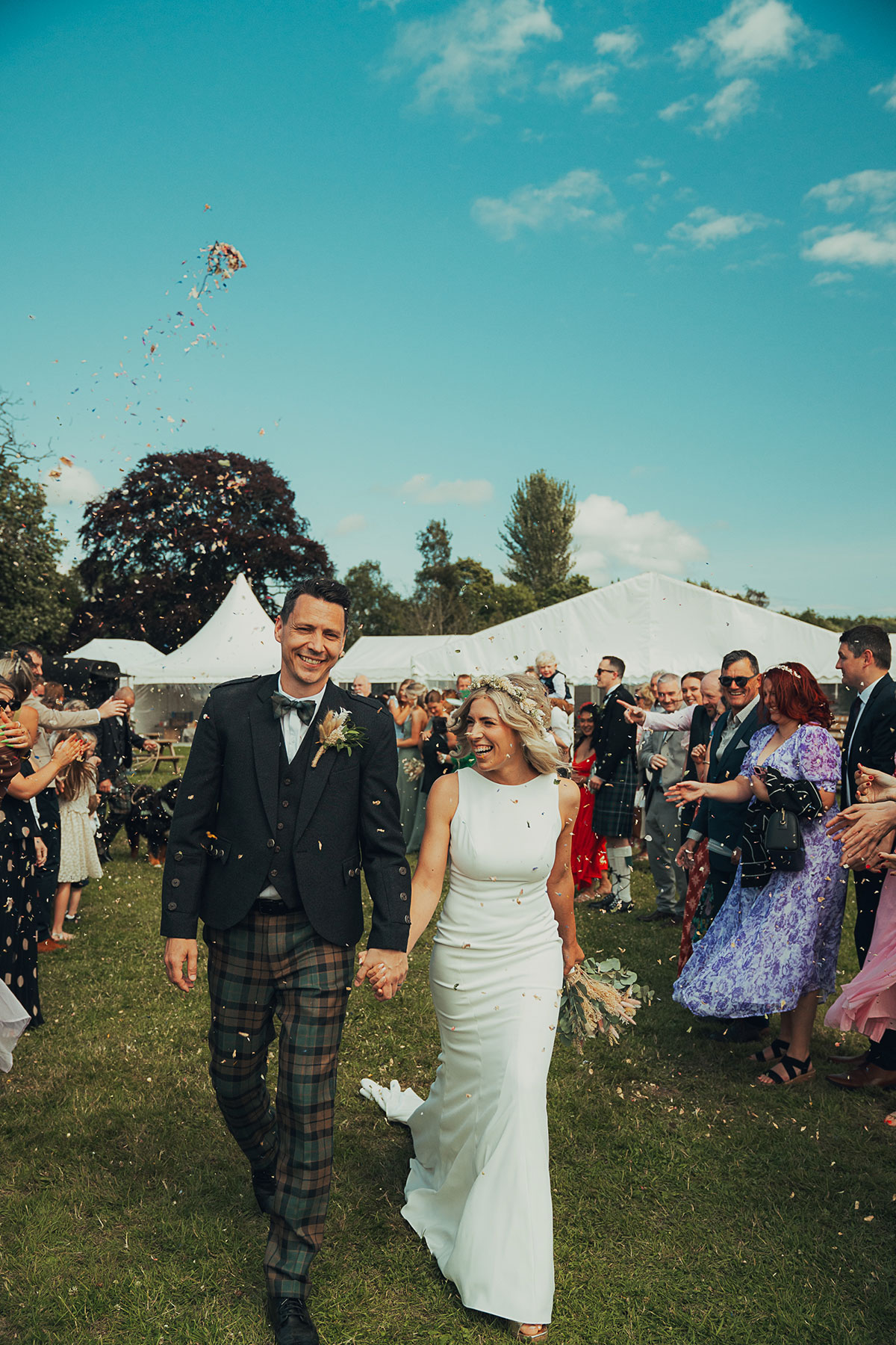 Bride and groom walk hand in hand through confetti outdoors, smiling as guests line either side near a marquee reception