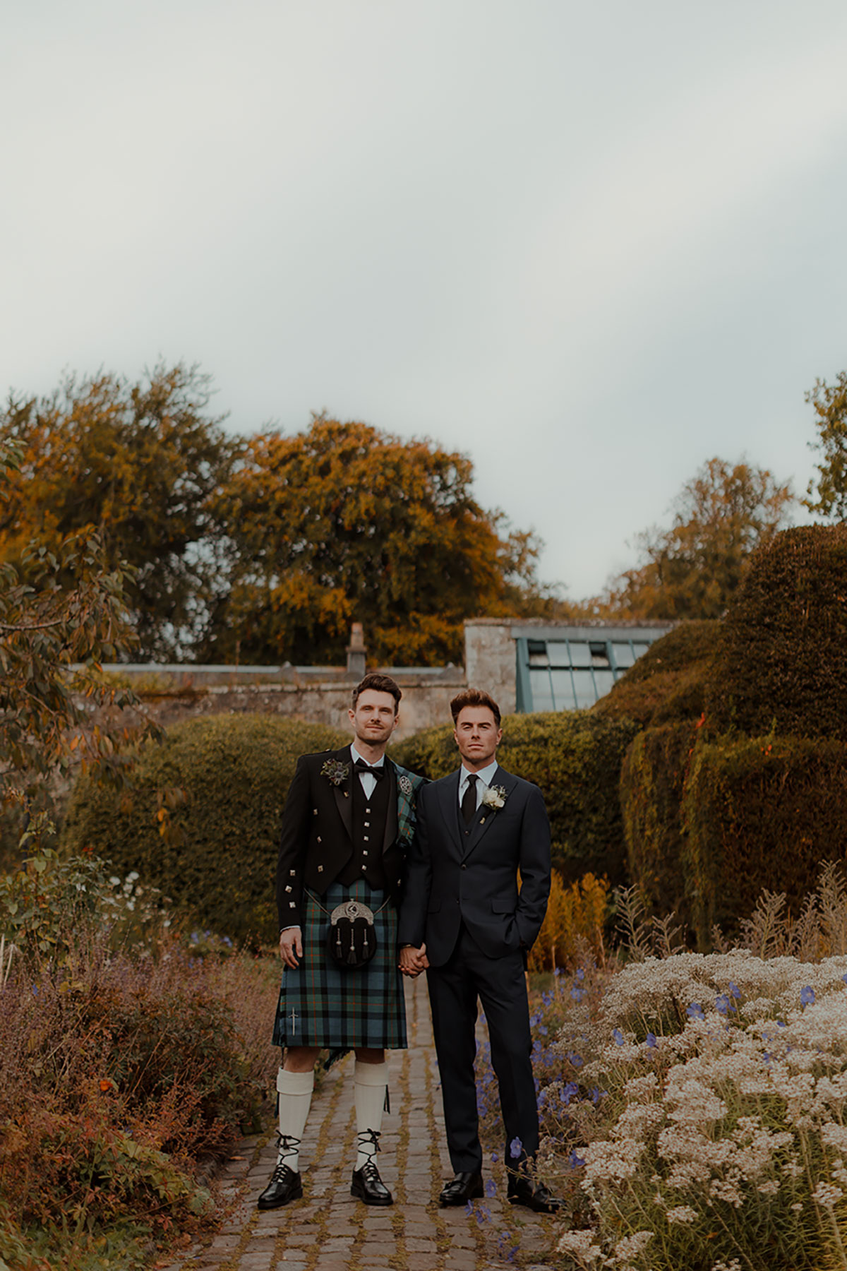 Two grooms stand hand in hand in the gardens at Newhall Estate, wearing a Gunn tartan kilt by MacGregor and MacDuff and a navy wedding suit