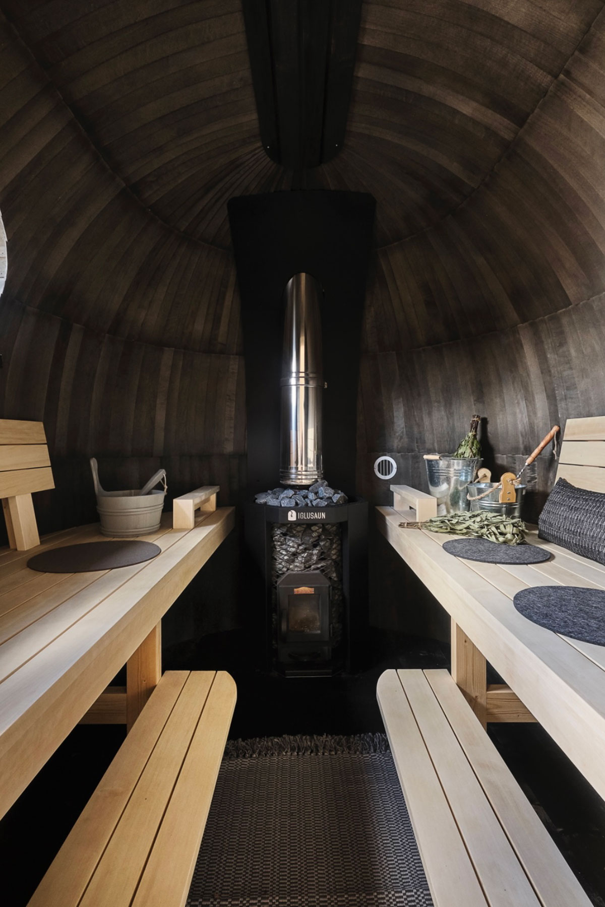 a black wooden sauna with the rocks and heat source in the centre and pale wooden benches along either side with small silver buckets of water sat on top