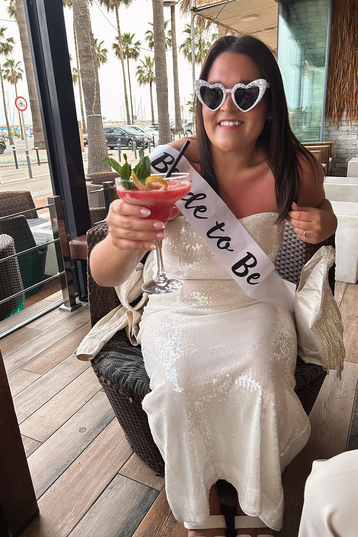 a bride to be holding a cocktail and wearing a sash and heart-shaped sun glasses