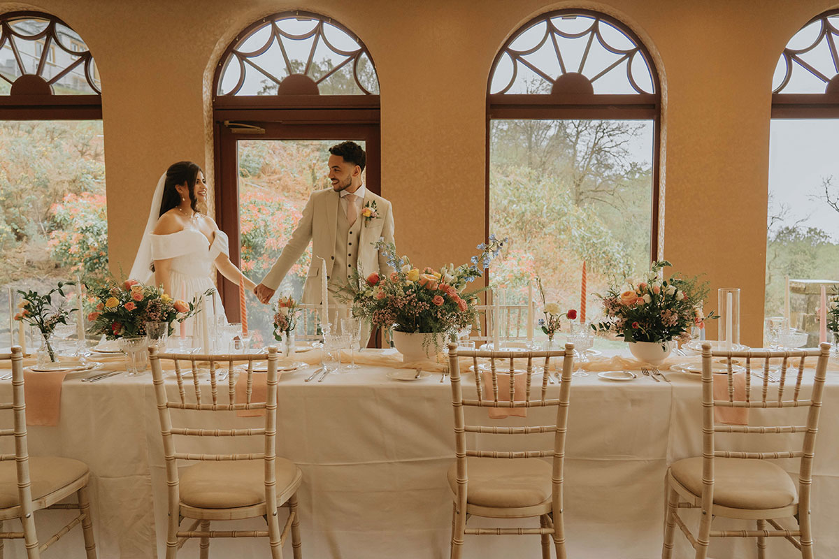 bride in off the shoulder dress and groom in tan suit with pale pink tie hold hands and walk along formally set dinner table with linens, flower arrangements and candlesticks