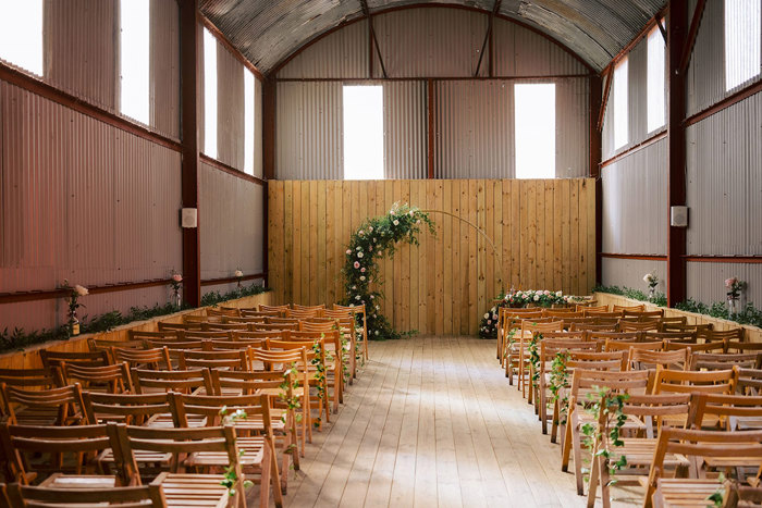The inside of a tall barn with rows of wooden chairs facing an arch with flowers and foliage across one half 