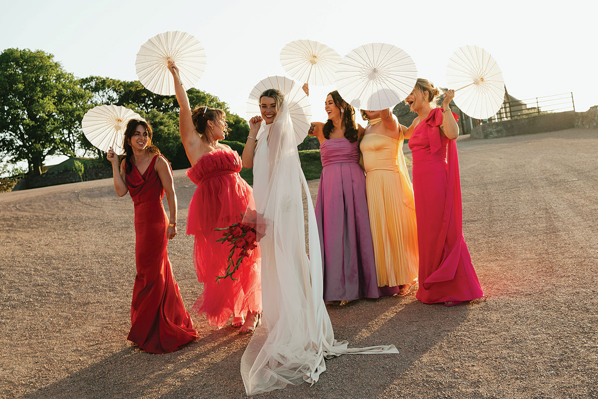 Bride with bridesmaids in colourful dresses holding white parasols at an outdoor summer wedding