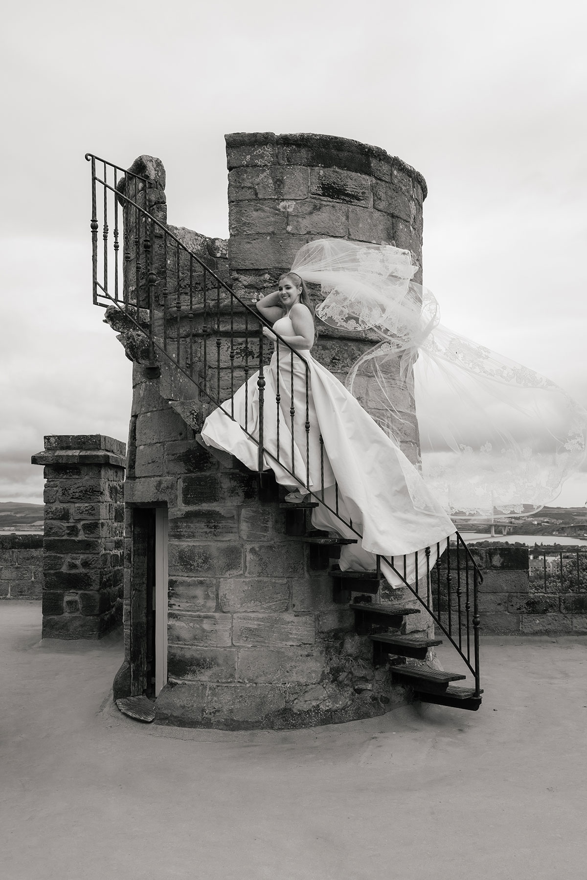 Bride stands on stone turret staircase at Dundas Castle, her veil blowing dramatically in the wind.