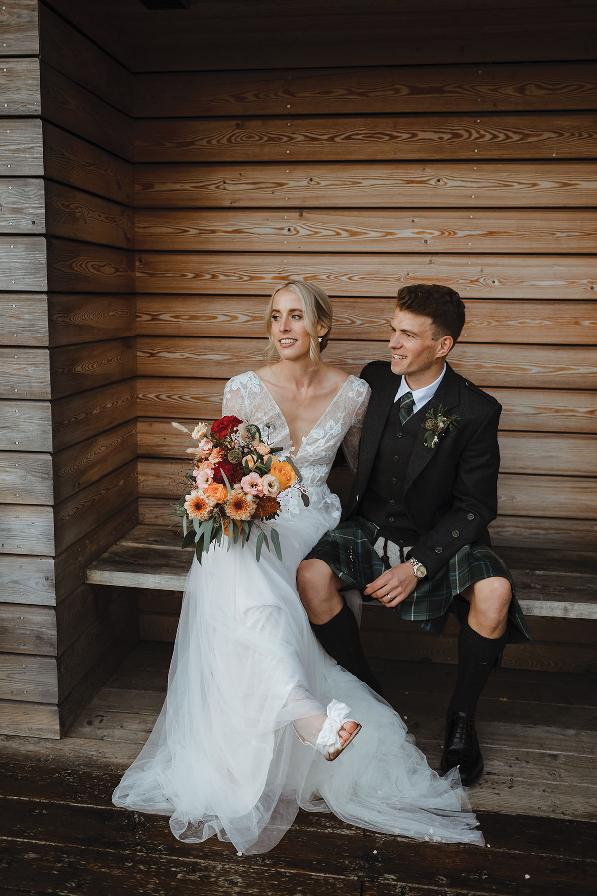 Bride and groom sitting on a wooden bench against timber cladding; the bride holds a bouquet of orange and red flowers and the groom wears a kilt and dark jacket