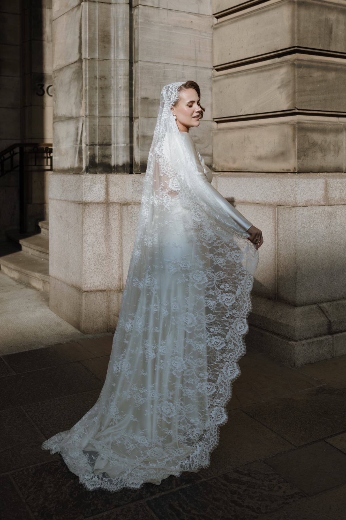 Bride posing in Glasgow city centre wearing a satin wedding dress and long floral lace veil.