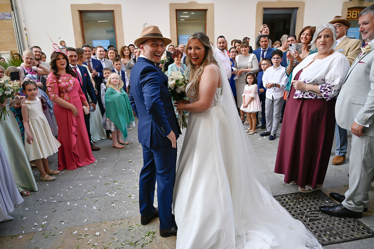 groom in blue suit and bride in ballgown with bouquet of white flowers stand in the middle of an empty space with flower petals on the ground and a ring of formally dressed people surrounding them