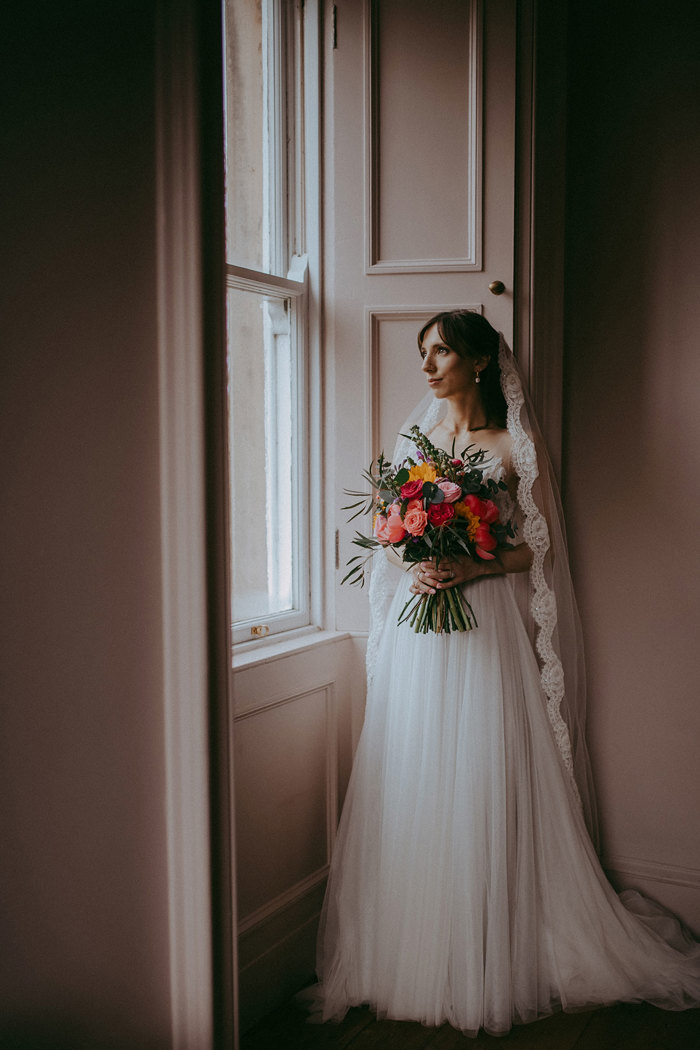 a bride posing by a window. She is wearing a lace-edged veil and holding a multi-coloured bouquet of flowers.