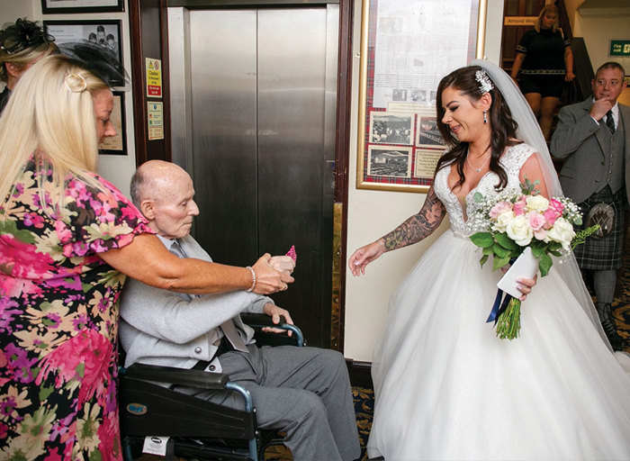 An elderly man in a wheelchair handing something to a bride in front of an elevator