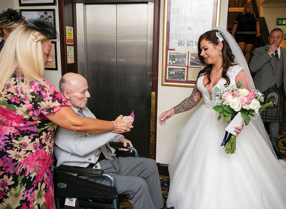 An elderly man in a wheelchair handing something to a bride in front of an elevator