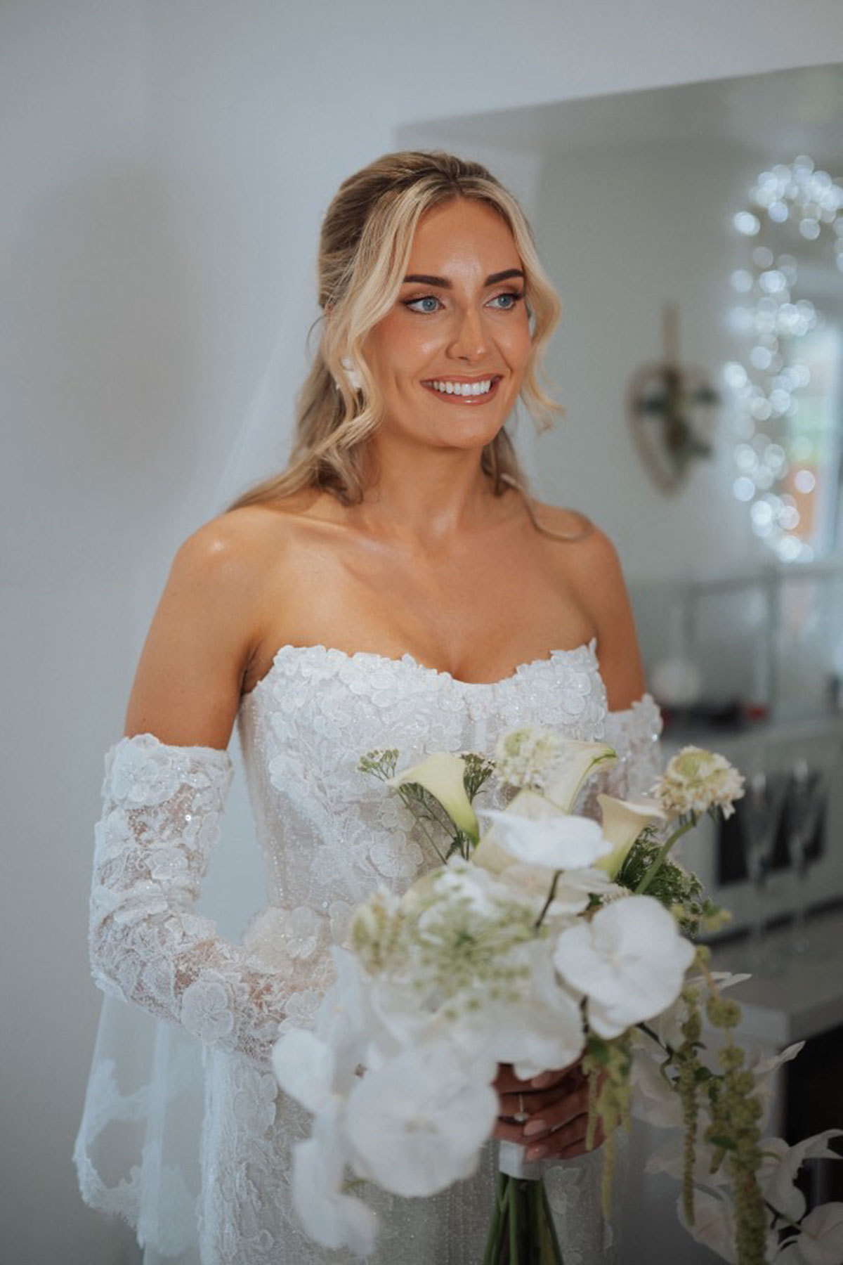 Bride holding white orchid wedding bouquet in lace off-the-shoulder gown during bridal preparations
