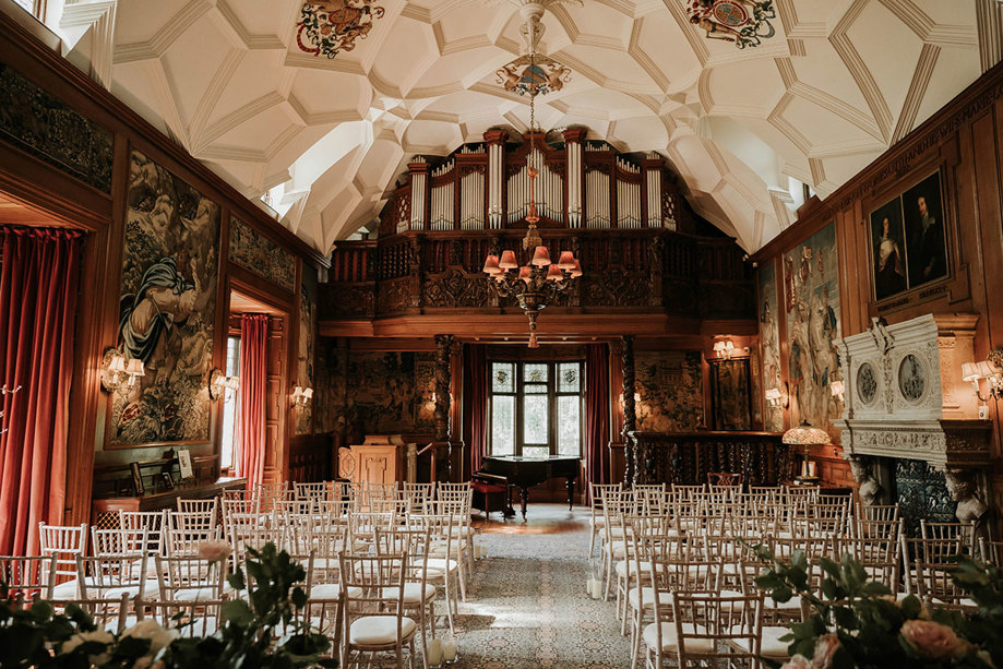 A grand hall with wooden panelling, white wooden chairs set out for a ceremony, an ornate white ceiling and dark green floral wallpaper
