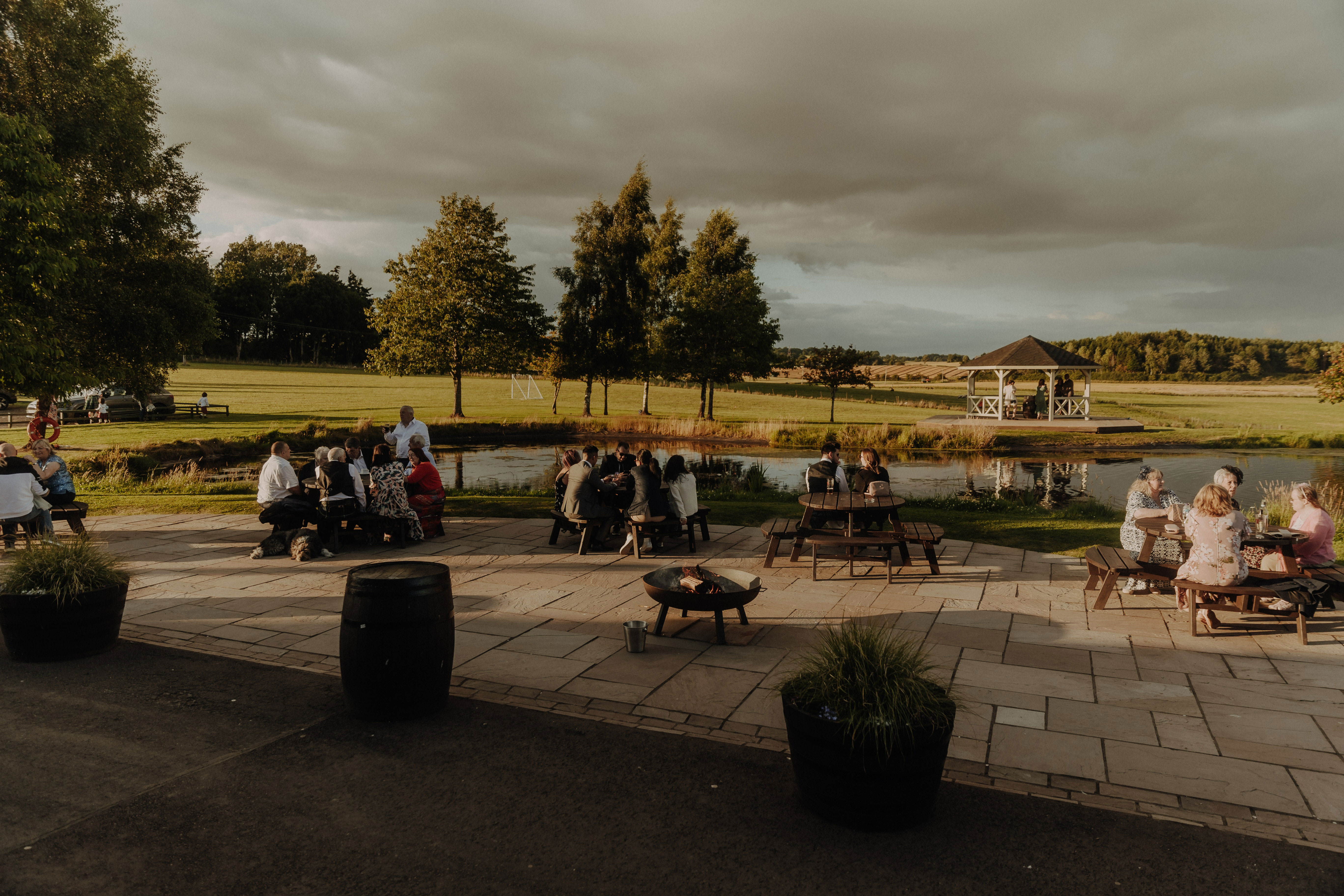 guests sit outside on communal benches talking to one another during wedding at bachilton barn as the sun goes down