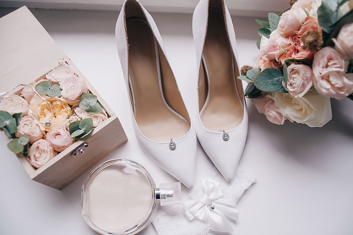 Pair of white court heels next to pink roses and a clear perfume bottle