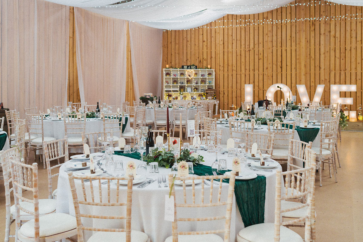 A bright reception space with round tables, green runners and large light-up ‘LOVE’ letters against a timber wall.