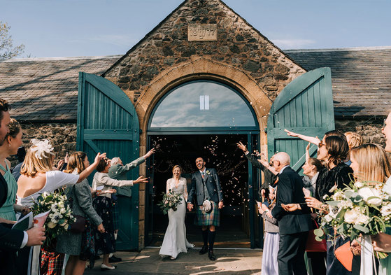 A bride and groom walking out of a byre as their guests throw confetti on them