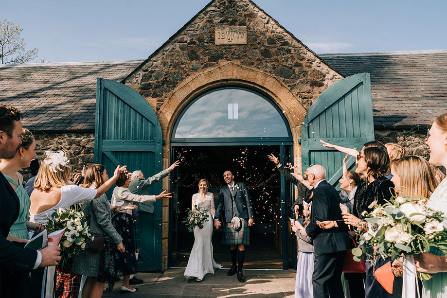 A bride and groom walking out of a byre as their guests throw confetti on them