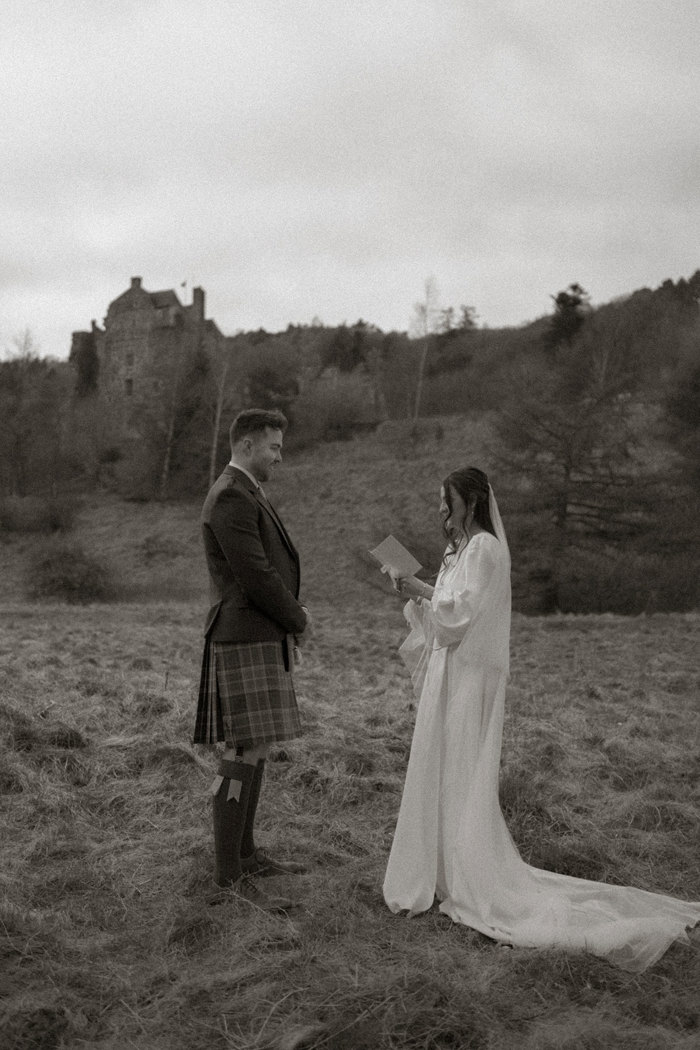 black and white image of bride in trailing satin dress with matching veil reading from a booklet, standing facing a groom in kilt outfit, stood outdoors on the grass with a small blurred castle in the background