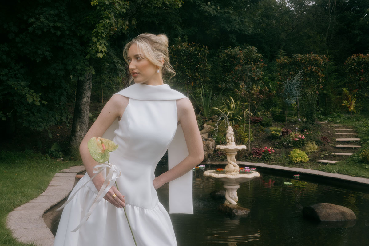 A bride in a modern white wedding gown stands beside a pond and stone fountain in a landscaped garden, holding a green anthurium flower.