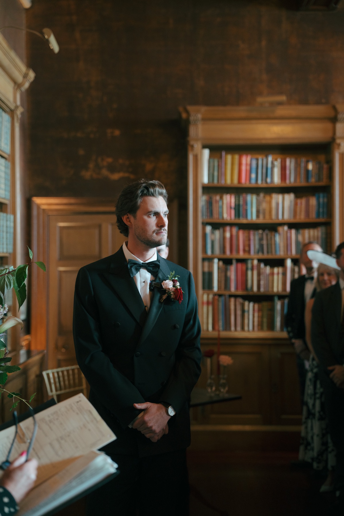 Groom Ryan waits for bride Nicole during their wedding ceremony in the library at Barnbougle Castle, wearing a black tuxedo and red boutonnière.