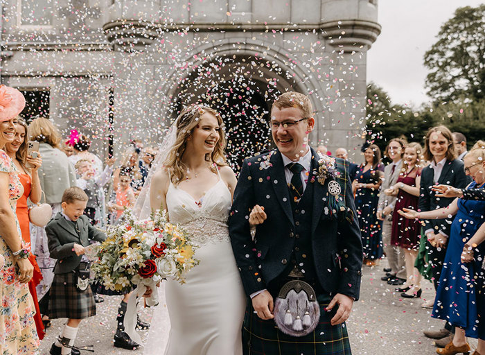 A bride and groom link arms and walk under colourful confetti 