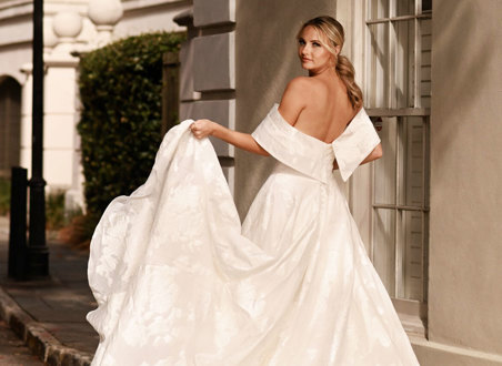 bride wearing large dramatic off-the-shoulder wedding dress looks back over her shoulder while holding up one corner of her large ballgown skirt