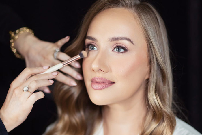 a hand reaches into frame with a makeup brush to add the finishing touches to a woman's makeup