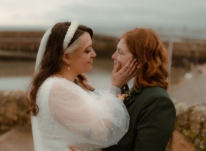 Two brides pose on their wedding day. One bride is wearing a pearl detail wedding dress and headband. She caresses the face of the second bride who is wearing a dark green suit. Boats in Crail Harbour are in the background